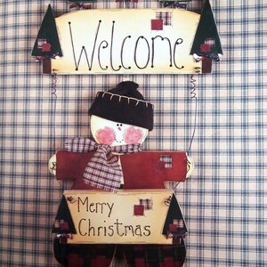 Puede incluir: Decoraci&oacute;n navide&ntilde;a de madera con forma de mu&ntilde;eco de nieve, sosteniendo un letrero que dice "Merry Christmas". El mu&ntilde;eco de nieve lleva un gorro negro, una bufanda a cuadros y un letrero de "Welcome" arriba. El fondo es un patr&oacute;n de cuadros azules y blancos.