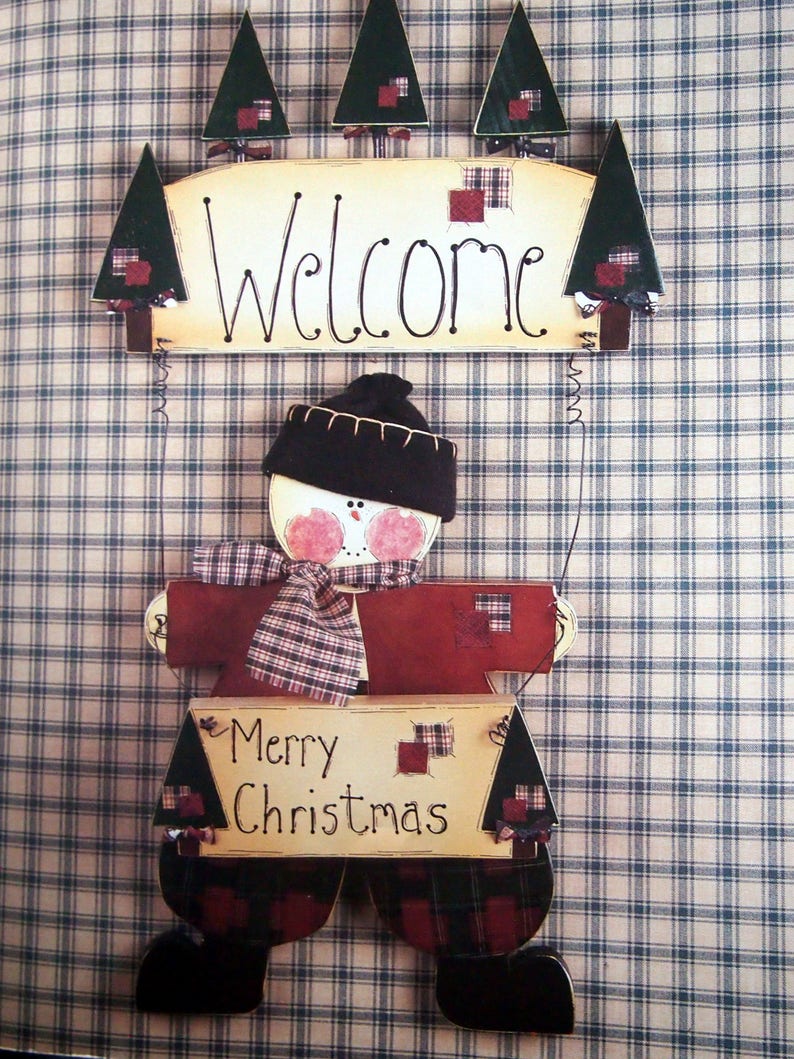 Puede incluir: Decoraci&oacute;n navide&ntilde;a de madera con forma de mu&ntilde;eco de nieve, sosteniendo un letrero que dice "Merry Christmas". El mu&ntilde;eco de nieve lleva un gorro negro, una bufanda a cuadros y un letrero de "Welcome" arriba. El fondo es un patr&oacute;n de cuadros azules y blancos.