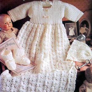 May include: A white crocheted baby gown, bonnet, and booties set. The gown has a lace pattern and is tied with a white ribbon. The set is laid out on a wooden surface with a certificate of baptism and a doll.