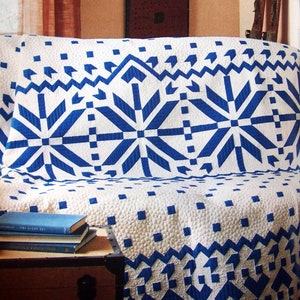 May include: A white and blue quilt draped over a sofa. The quilt features a repeating geometric pattern of blue starbursts and zigzags on a white background. The quilt is made of a textured fabric and is folded over the sofa.