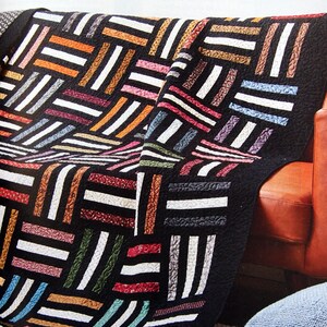 May include: A colourful quilt with a geometric pattern of black, white, red, orange, brown, blue, and green stripes. The quilt is draped over a red leather sofa with a blue throw pillow.