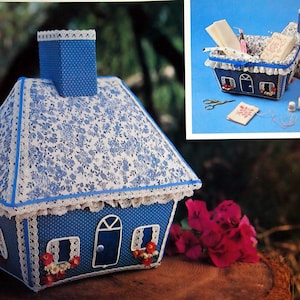 Puede incluir: Una caja de costura en forma de casa con un estampado floral azul y blanco y un borde de encaje blanco. La caja está abierta y llena de artículos de costura, incluyendo hilo, agujas y tijeras. La caja está sentada sobre una superficie de madera.