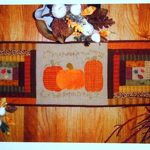 May include: A brown and beige quilted table runner with a stitched design of three pumpkins and the text "Pumpkin Patch".