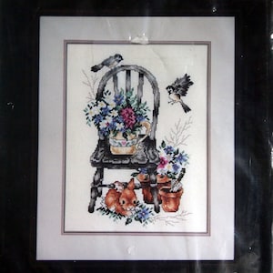 May include: Counted cross stitch pattern titled "The Beauty of Spring" by Janlynn. The pattern features a black chair with a flower pot on the seat, a small rabbit, and two birds. The finished size is 27.9 cm by 35.6 cm.