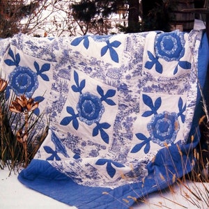 May include: A blue and white quilt with a floral pattern. The quilt is made of cotton fabric and has a blue border. The quilt is folded and lying on a snowy surface.