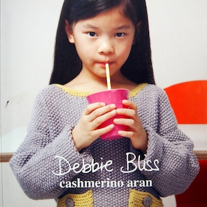 May include: A young person wearing a gray sweater with yellow trim and accents, holding a pink cup with a straw. The text "Debbie Bliss cashmerino aran" is visible on the image.