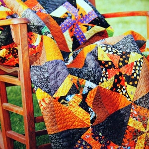 May include: A colorful patchwork quilt with a Halloween theme, featuring orange, black, and purple fabrics with various patterns, including stars, polka dots, and swirls. The quilt is draped over a wooden rocking chair.