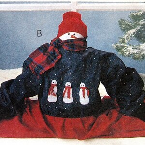 May include: A snowman wearing a blue sweater with white snowflakes, a red hat, and a red and black plaid scarf. The snowman has three smaller snowmen on its sweater. The snowman is sitting with its legs spread out and wearing black shoes.