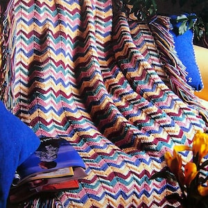 May include: A multi-coloured crocheted throw blanket with fringe on the edges. The blanket is draped over a sofa with a blue cushion and a stack of magazines.