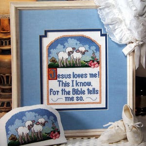 May include: A framed cross-stitch design featuring two white lambs in a field with blue sky and clouds. The text reads "Jesus loves me! This I know. For the Bible tells me so."