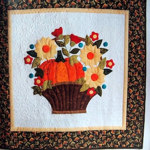 May include: A quilted wall hanging with a brown basket filled with orange pumpkins and yellow flowers. The basket is surrounded by green leaves and red flowers. The background is a white fabric with a quilted pattern.