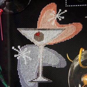 May include: Cross-stitch design of a martini glass with an olive, set against a black background. The glass is white with a pink and white accent, and a small decorative element. Cocktail theme.