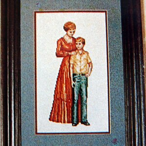 Puede incluir: Un bordado de punto de cruz enmarcado que representa a una mujer con un vestido rojo y un ni&ntilde;o de pie a su lado. La mujer est&aacute; sosteniendo la mano del ni&ntilde;o. El bordado est&aacute; sobre un fondo azul.