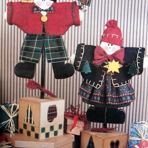 Puede incluir: Decoraciones de mu&ntilde;ecos de nieve navide&ntilde;os hechos a mano. Un mu&ntilde;eco de nieve lleva un abrigo rojo y pantalones a cuadros, el otro un abrigo y una falda negros. Ambos tienen sombreros negros y est&aacute;n sobre bases de bloques de madera. Regalos en primer plano.