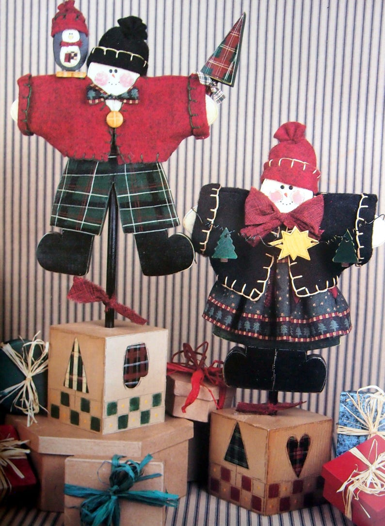 Puede incluir: Decoraciones de mu&ntilde;ecos de nieve navide&ntilde;os hechos a mano. Un mu&ntilde;eco de nieve lleva un abrigo rojo y pantalones a cuadros, el otro un abrigo y una falda negros. Ambos tienen sombreros negros y est&aacute;n sobre bases de bloques de madera. Regalos en primer plano.