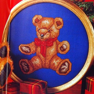 May include: A brown teddy bear with a red bow sits in a gold frame on a blue background. The teddy bear is a cross-stitch design.