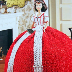 May include: A doll in a red and white crocheted dress with a matching scarf. The dress features a full skirt and a white bodice with red detailing. The doll holds a small book. The image is labelled "The Southern Belle Collection Holiday Dress."