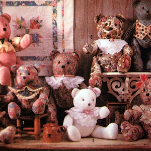May include: A collection of handmade teddy bears in various sizes and patterns. The bears are made from fabrics in shades of pink, floral prints, and checkered patterns. Some bears have decorative collars and bows. The background includes a quilt and a small shelf.