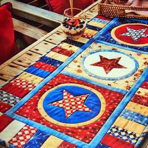 Puede incluir: Un camino de mesa de tela rectangular con un diseño de patchwork patriótico en colores rojo, blanco y azul. El camino muestra motivos de estrellas acolchadas dentro de marcos circulares, alternando con patrones de rayas y cuadros.
