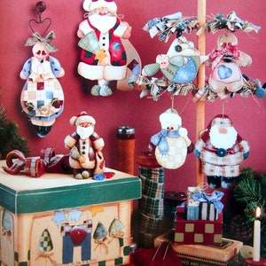 May include: A collection of handcrafted holiday ornaments featuring Santa Claus, snowmen, and figures with fabric accents, hanging from wire loops and a wooden stand. A painted wooden box with a green lid and fabric-wrapped spools are also present.