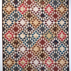 Op de afbeelding: Een grote patchwork quilt met een complex geometrisch ontwerp. De quilt toont een herhalend patroon van ruiten en driehoeken in rood, blauw, crème en bruin. De rand is donkergrijs. Een handgemaakt textiel.