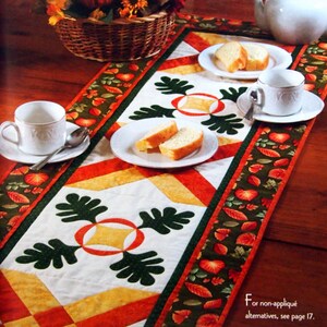 May include: A white table runner with a patchwork design featuring orange, yellow, and green squares and leaves. The runner is decorated with a central motif of three green oak leaves with a red and yellow circle in the center. The runner is set on a wooden table with a basket of fall decor, a teapot, and two plates of pastries.