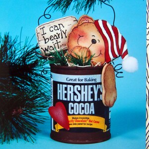 May include: A decorative Hershey's Cocoa can with a bear wearing a red and white striped hat. The can has a red heart and a gingerbread cookie attached. Text on the can reads "Great for Baking" and "HERSHEY'S COCOA". A sign says "I can bearly wait..."