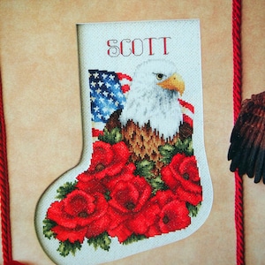 May include: A white stocking with a red, white, and blue American flag, a bald eagle, and red poppies. The stocking is personalized with the name "SCOTT".