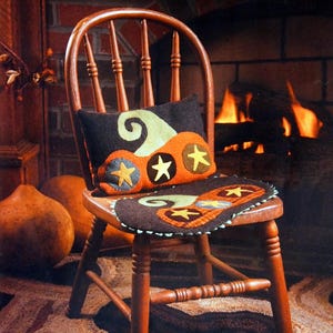Puede incluir: Un cojín rectangular de fieltro marrón oscuro y un cojín de asiento ovalado a juego con una aplicación de calabaza naranja, espirales verde claro y estrellas amarillas. El conjunto se exhibe sobre una silla de madera.