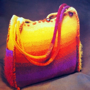 May include: A large, colorful knitted tote bag with a purple, orange, and yellow ombre design. The bag has a single strap and a small, round, red button.