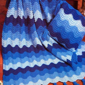 May include: A handmade crochet blanket and cushion set in shades of blue. The blanket has a wavy pattern with varying shades of blue. The cushion matches the blanket's design. Blue tassels decorate the blanket's edge.