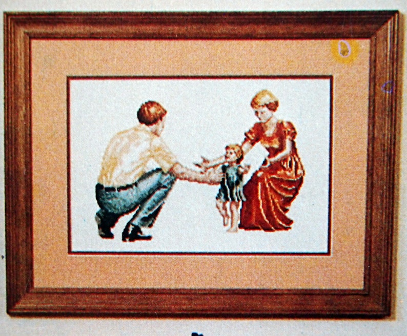 Puede incluir: Un bordado de punto de cruz enmarcado que representa a una familia de tres. Un hombre con una camisa amarilla y pantalones vaqueros azules est&aacute; tendi&eacute;ndole la mano a un ni&ntilde;o peque&ntilde;o con un vestido verde. Una mujer con un vestido rojo est&aacute; sosteniendo la mano del ni&ntilde;o. El bordado est&aacute; sobre un fondo beige.