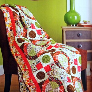 Op de afbeelding: Een kleurrijk patchwork-deken met cirkels, vierkanten en bloemmotieven in tinten rood, groen, bruin en wit. Het deken is over een bruine fauteuil gelegd.