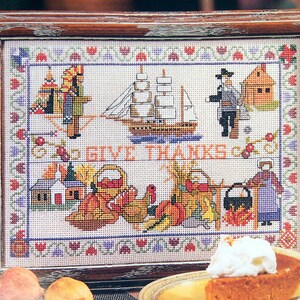 May include: A framed cross-stitch sampler featuring the text GIVE THANKS, depicting a sailing ship, a teepee, a cabin, a person in a hat, a turkey, a pumpkin, a cooking pot over a fire, and a person in a bonnet, all bordered by floral motifs.