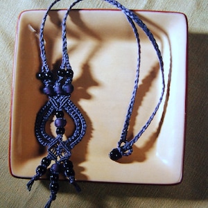 Peut inclure: Un collier en macramé de couleur violet foncé et noir avec des perles. Le collier est posé sur une assiette carrée de couleur marron clair.