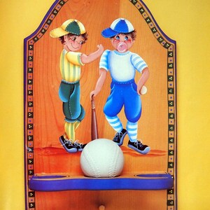 May include: A wooden wall decor with a blue and yellow border. Two cartoon boys are playing baseball. One boy is wearing a blue hat and a white shirt with blue stripes. The other boy is wearing a green hat and a yellow and white striped shirt. A white baseball is on a shelf below the boys. The text "LOOK AT THE MUSCLE pages 17, 18" is at the bottom of the decor.