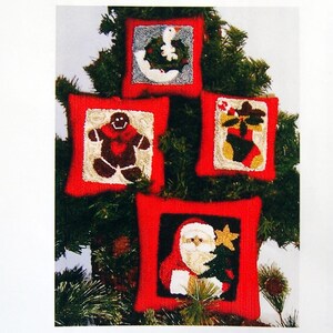 May include: Four Christmas-themed decorative pillows with red borders are displayed on a Christmas tree. The pillows feature images of a goose with a wreath, a gingerbread man, a Christmas stocking, and Santa Claus holding a tree and star.