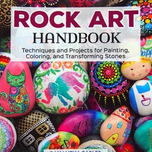 May include: A vibrant collection of painted rocks, showcasing various designs and techniques. The image features a book titled "ROCK ART HANDBOOK" with the author's name, Samantha Sarles. The rocks are decorated with colorful patterns, animals, and other creative motifs.
