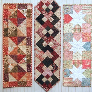 May include: Three quilted table runners in various designs. The left runner has a patchwork design with triangles in red, orange, and brown. The centre runner has a geometric pattern in red, black, and cream. The right runner has a star pattern.