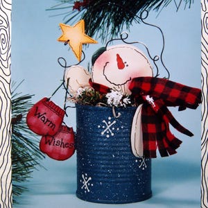 May include: A festive snowman decoration in a blue tin can. The snowman features a painted face, a red and black plaid scarf, and red mittens with the words "Warm Wishes." A yellow star and greenery complete the winter scene.