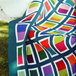 May include: A colorful quilt with a teal background and a geometric pattern of rectangles in various colors, including red, purple, green, and yellow. The quilt is folded over a white surface, with a green lawn in the background.