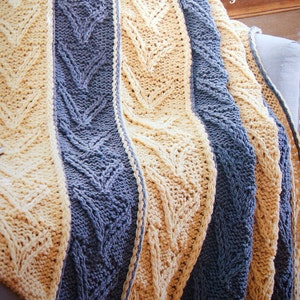 Op de afbeelding: Een gebreide deken met een kabelbreipatroon in beige en blauwe tinten. De deken is gevouwen en over een stoel gelegd.