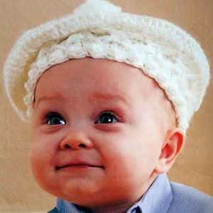 May include: A white crocheted beret with a small pompom on top. The beret is worn by a baby wearing a light blue shirt.