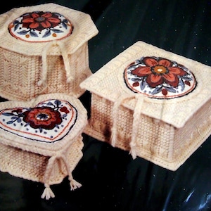 May include: Three crocheted boxes with floral embroidery. The boxes are in the shape of a square, octagon, and heart. The boxes are cream colored with a white crocheted design. The flowers are red, orange, and brown.