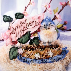 May include: A bluebird with a pink and white nest, sitting on a nest of straw. A pink banner with the words "To Mothers" is behind the bird.