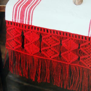 May include: A close-up of a white tablecloth with red stripes and a fringed diamond border. The border is a vibrant red, featuring geometric patterns and long fringe. A small dish with walnuts sits on the table.