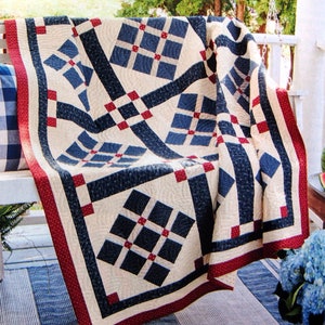 May include: A red, white and blue quilt with a geometric pattern. The quilt is made of cotton fabric and is quilted with a diamond pattern. The quilt is folded and draped over a white porch swing.
