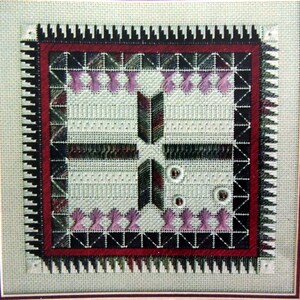 May include: A close-up of a cross-stitch embroidery design featuring a geometric pattern with black, white, red, and green thread. The design is framed with a black and red border.