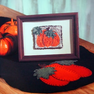 May include: A framed needlepoint picture of three orange pumpkins with green leaves on a white background. The frame is brown wood and the picture is sitting on a black table runner with two orange felt pumpkins with green leaves.