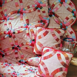 May include: A set of three pink and red quilted throw cushions with a double ring design. The cushions are arranged on a pink and red quilted throw blanket with a double ring design. There are three red apples in the foreground.